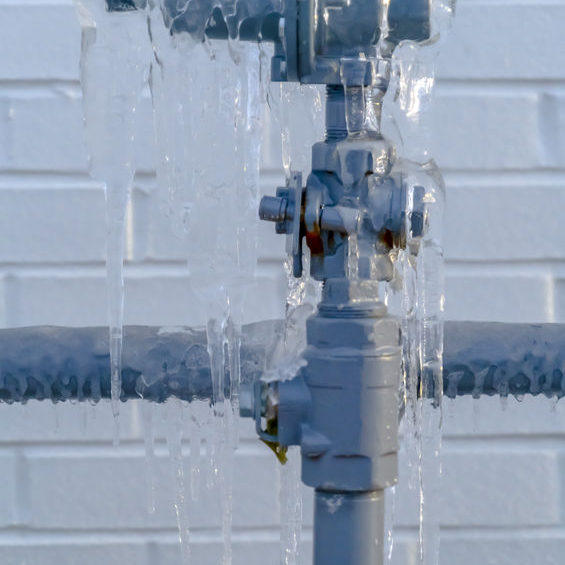 Close up of a gray metal pipe covered ice with against a white brick wall. View outside a building in Eagle Mountain, Utah during winter.