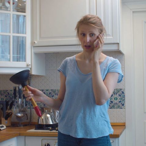 Confused woman in the kitchen with a plunger in her hands. She is calling by smart phone for advice. Kitchen sink clogging. Plumbing in need of repair. Housekeeping